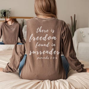 May include: A brown long-sleeve shirt with the text "there is freedom found in surrender" and "proverbs 3:5-6" printed on the back in white. The shirt also has a small heart design on the front.