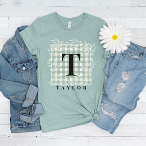 May include: A light green t-shirt with a white and green gingham pattern and a black letter T in the centre. The t-shirt has a floral design around the gingham pattern and the name "TAYLOR" printed below the gingham pattern.