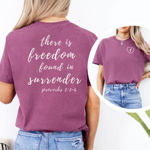 May include: A plum-colored short-sleeve t-shirt featuring white cursive text on the back that reads there is freedom found in surrender proverbs 3:5-6. The front of the shirt displays a small white heart graphic with a cross inside on the chest.
