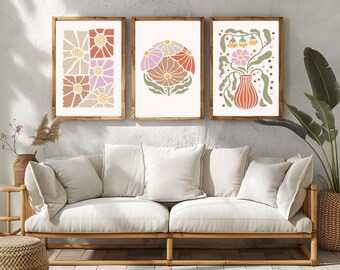 Op de afbeelding: Drie ingelijste prints met abstracte bloemenmotieven in roze, oranje en groene tinten. De prints hangen aan een witte muur boven een lichtbruine bank.