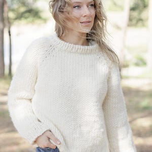 May include: A chunky knit sweater in an off-white cream color featuring a round neckline, long sleeves with raglan-style seams, and ribbed cuffs and hem. The garment is crafted with a thick, textured yarn in a classic stockinette stitch pattern.