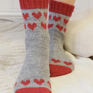 Könnte beinhalten: Ein Paar wadenhohe Strickstrümpfe mit einer meliert grauen Basis und einem wiederkehrenden Muster aus kleinen roten Herzen. Die Socken haben ein geripptes rotes Bündchen am oberen Rand und eine einfarbig rote Spitze mit sichtbarer Strickstruktur.