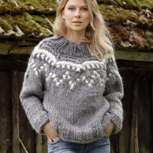 May include: A chunky knit sweater in heather gray with a white zigzag pattern around the yoke and neckline. The sweater features long sleeves, a ribbed crew neck, and a thick, textured wool construction paired with light blue denim jeans.