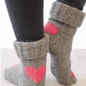 May include: Gray knit socks with a large pink heart design on the side. The socks have a folded-over cuff and are worn with black pants. The socks appear to be handmade and are a cozy, warm style.