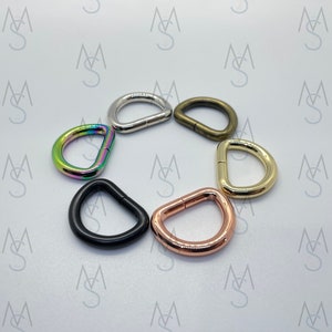 May include: Seven D-shaped metal rings in different colors, including silver, gold, black, rose gold, and iridescent. The rings are arranged in a circle on a white background.