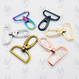 Może przedstawiać: Pięć metalowych karabińczyków w różnych kolorach. Karabińczyki są w kolorze srebrnym, złotym, różowym złocie, czarnym i tęczowym.