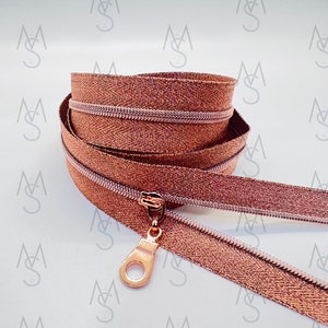 May include: A close-up of a rose gold zipper with a rose gold slider. The zipper is made of a metallic fabric with a subtle shimmer.
