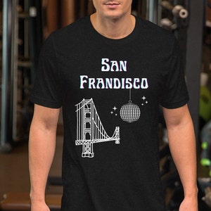 Puede incluir: Camiseta negra con un gr&aacute;fico blanco del Golden Gate Bridge, una bola de discoteca y el texto "San Frandisco". El dise&ntilde;o tiene un aire retro y vintage. La camiseta est&aacute; hecha de un material suave.