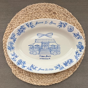 May include: An oval white ceramic platter featuring blue line art illustrations. The center depicts a barn with a bow above it, labeled "Cripps Barn, Cotswolds, UK". The rim includes floral motifs, a dog face, and the text "Becca & Ben" and "June 10, 2026".