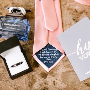 May include: A pink tie with a navy blue pocket square that says "I will love you for all the days of my life. You are my forever. Love, your Gri" in white text. A small gray card with the text "his vows" in white cursive writing is on the table next to the tie.