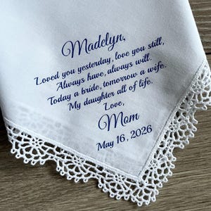 Może przedstawiać: Biała chusteczka z delikatnym koronkowym obszyciem. Chusteczka zawiera niebieski napis z tekstem "Madelyn, Loved you yesterday, love you still...Love, Mom May 16, 2026". Sentimentalna pamiątka.