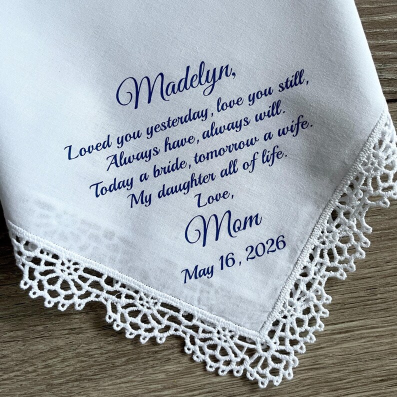 Może przedstawiać: Biała chusteczka z delikatnym koronkowym obszyciem. Chusteczka zawiera niebieski napis z tekstem "Madelyn, Loved you yesterday, love you still...Love, Mom May 16, 2026". Sentimentalna pamiątka.
