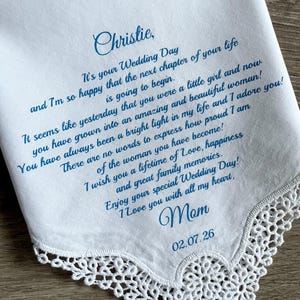 May include: White handkerchief with blue script. The text reads "Christie, It's your Wedding Day...I Love you with all my heart, Mom 02.07.26". The edge of the handkerchief has a delicate lace trim.