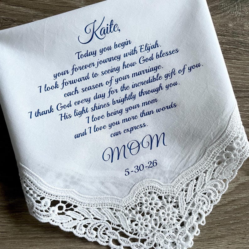 May include: White handkerchief with delicate lace trim. The handkerchief has blue script that reads "Kaite" and a heartfelt message from "Mom" with the date 5-30-26. A sentimental keepsake.