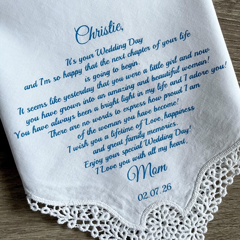 Może przedstawiać: Biała chusteczka z niebieskim pismem. Tekst brzmi: "Christie, To Tw&oacute;j dzień ślubu... Kocham Cię całym sercem, Mamo 02.07.26". Krawędź chusteczki ma delikatne koronkowe wykończenie.