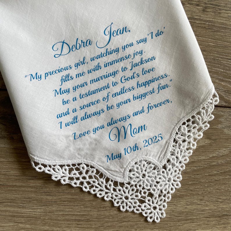 May include: White handkerchief with delicate lace trim and blue script. The text reads "Debra Jean" followed by a heartfelt message from "Mom" and the date "May 10th, 2025". A sentimental keepsake.
