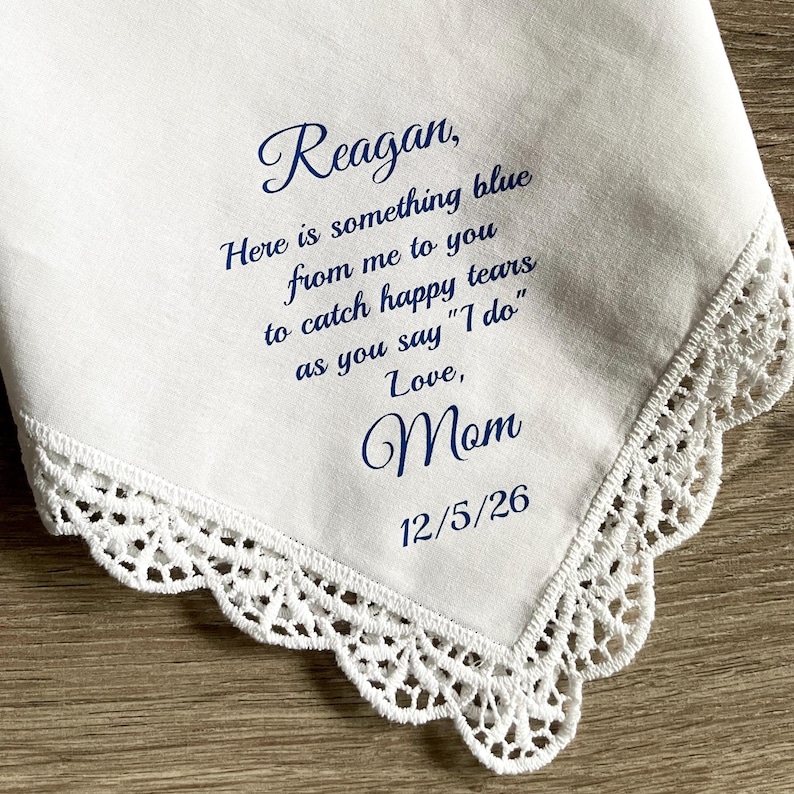 May include: White handkerchief with a delicate lace trim. The handkerchief has blue text that reads "Reagan, Here is something blue from me to you to catch happy tears as you say "I do" Love, Mom 12/5/26."
