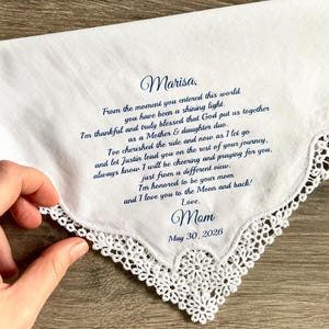 May include: White handkerchief with a delicate lace trim. The handkerchief has a handwritten message in navy blue ink, including the name "Marisa" and the date "May 30, 2026". The message is from "Mom".