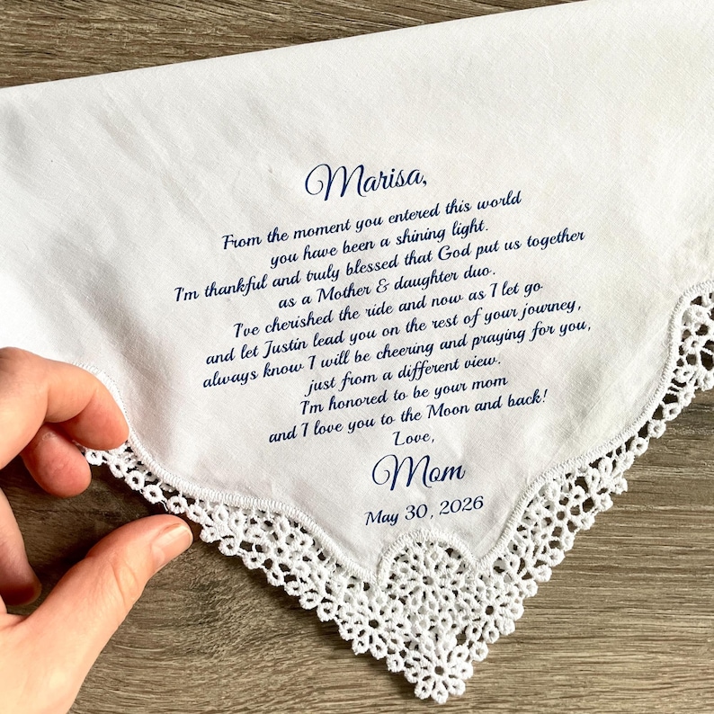 May include: White handkerchief with a delicate lace trim. The handkerchief has a handwritten message in navy blue ink, including the name "Marisa" and the date "May 30, 2026". The message is from "Mom".