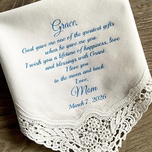 May include: White handkerchief with blue script. The text reads "Grace, God gave me one of the greatest gifts when he gave me you. I wish you a lifetime of happiness, love and blessings with Grant. I love you to the moon and back. Love, Mom March 7, 2026." Features a lace trim.