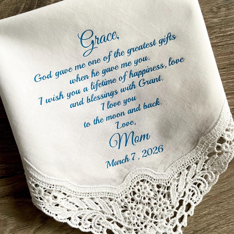 May include: White handkerchief with blue script. The text reads "Grace, God gave me one of the greatest gifts when he gave me you. I wish you a lifetime of happiness, love and blessings with Grant. I love you to the moon and back. Love, Mom March 7, 2026." Features a lace trim.