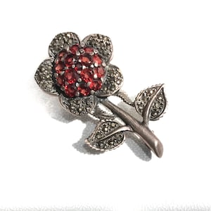 Puede incluir: Un broche de plata con forma de flor con piedras preciosas rojas y detalles de marcasita.