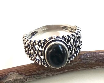 May include: A silver ring with an oval, black gemstone center. The ring has a detailed, openwork design around the gemstone and band. The ring is displayed on a small, brown twig against a white background.