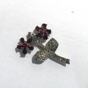 Puede incluir: Un broche de plata con dos acentos de flores rojas y un tallo de marcasita.