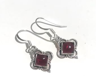 May include: A pair of silver earrings with a square, deep red gemstone set in a decorative, four-leaf clover-shaped frame. The earrings have a hook closure and are displayed on a white background.
