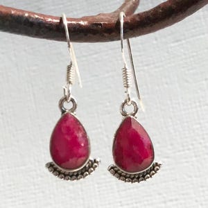 Puede incluir: Un par de pendientes colgantes de plata con piedras preciosas rojas oscuras en forma de lágrima. Las piedras están engastadas en un bisel de plata con un borde decorativo de cuentas. Los pendientes cuelgan de ganchos de plata.
