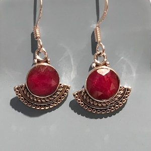 May include: A pair of silver earrings with a large, round, deep red gemstone at the center. The gemstone is set in a silver bezel and is surrounded by a decorative, fan-shaped silver design. The earrings have a hook closure.