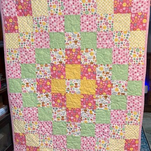 May include: A patchwork quilt with a pink border. The quilt features a grid pattern of squares in pink, yellow, and green, with floral and polka dot patterns. The quilt is likely handmade.