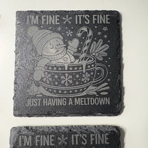 May include: Two square, dark gray slate coasters with etched designs. One coaster features a snowman in a mug with the text "I'M FINE IT'S FINE JUST HAVING A MELTDOWN". The other coaster has the same text at the top.