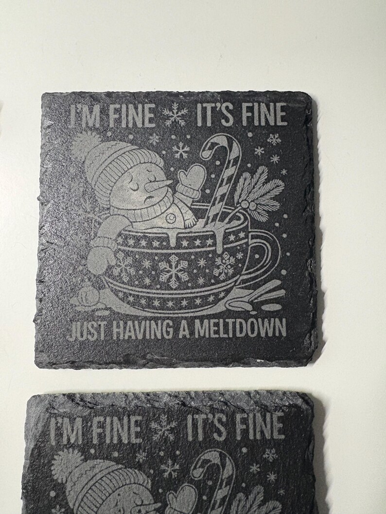 May include: Two square, dark gray slate coasters with etched designs. One coaster features a snowman in a mug with the text "I'M FINE IT'S FINE JUST HAVING A MELTDOWN". The other coaster has the same text at the top.