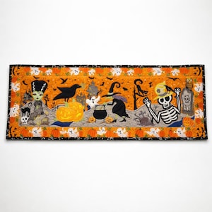 May include: A rectangular Halloween-themed table runner featuring a vibrant orange background with a quilted pattern. The design includes embroidered figures of a skeleton, a witch, a black cat, a crow, pumpkins, ghosts, and a haunted house.