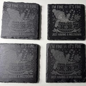 May include: Four square slate coasters with etched designs. Each coaster features a snowman in a teacup with the phrases "I'M FINE IT'S FINE" and "JUST HAVING A MELTDOWN". The coasters have a dark gray color.
