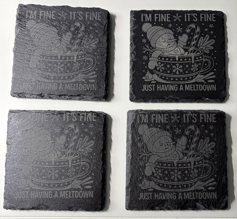 May include: Four square slate coasters with etched designs. Each coaster features a snowman in a teacup with the phrases "I'M FINE IT'S FINE" and "JUST HAVING A MELTDOWN". The coasters have a dark gray color.