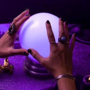 May include: A glowing, white crystal ball sits on a purple surface, with two hands framing it. The hands are adorned with rings and bracelets. A small candle and other decorative items are also visible.