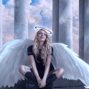 May include: A woman with long blonde hair, wearing a black top and boots, sits with large white feathered wings spread behind her. A halo is above her head. The background features white columns and a cloudy sky.