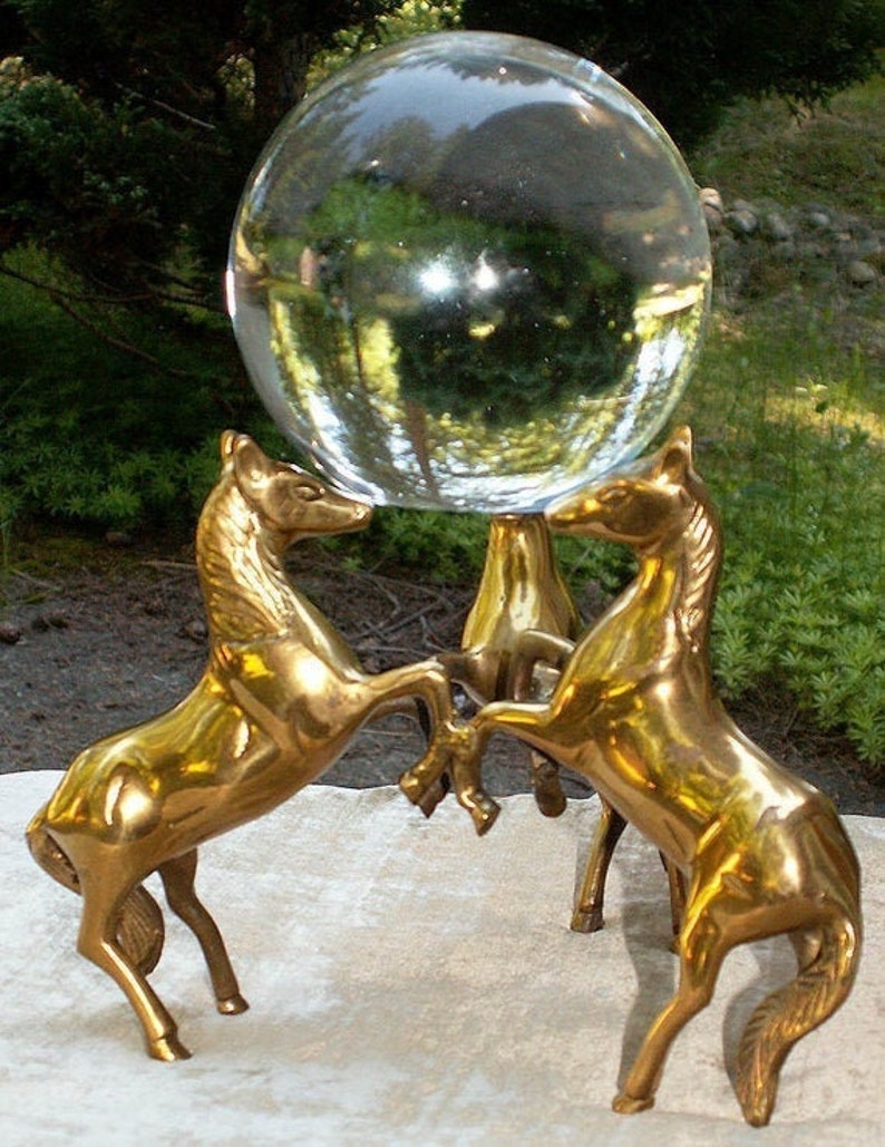 May include: A clear glass gazing ball rests on a brass stand supported by three rearing horse sculptures. The horses are golden in color, and the sphere reflects the surrounding greenery. This decorative piece is suitable for home decor.
