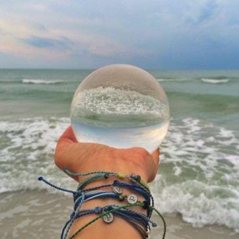 May include: A clear glass sphere held in front of a beach scene, reflecting the ocean and sky. Several blue and green bracelets are worn on the wrist. The ocean waves are visible in the background.