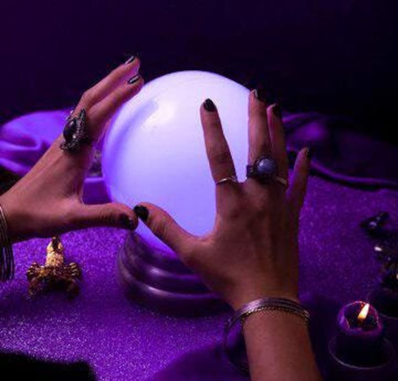 May include: A glowing, white crystal ball sits on a purple surface, with two hands framing it. The hands are adorned with rings and bracelets. A small candle and other decorative items are also visible.