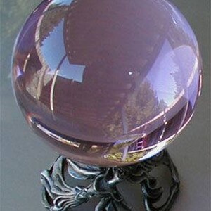 May include: A large, clear, lavender-colored crystal ball sits atop an ornate, silver-colored metal stand. The stand features a detailed, sculpted design. The crystal ball reflects the surrounding environment, creating a mirrored effect.
