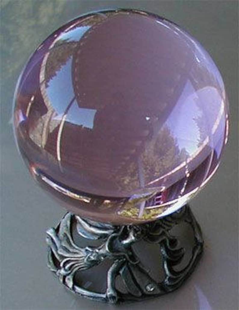 May include: A large, clear, lavender-colored crystal ball sits atop an ornate, silver-colored metal stand. The stand features a detailed, sculpted design. The crystal ball reflects the surrounding environment, creating a mirrored effect.