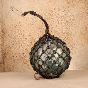 May include: A spherical glass fishing float encased in a dark brown knotted rope net. The netting extends upward into a thick rope handle, which is attached to a silver-colored metal safety pin at the top. The glass orb has a translucent blue-green tint.
