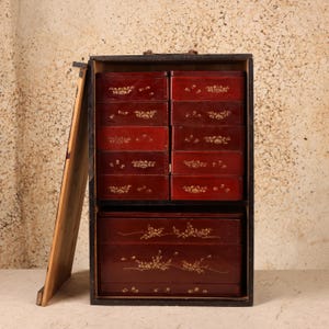 May include: A dark-colored wooden storage cabinet with a hinged door, featuring ten small red drawers arranged in two columns above one large red drawer. Each drawer front is decorated with gold-colored floral patterns and motifs.