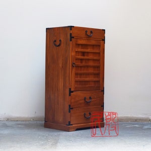May include: A wooden cabinet with a dark brown finish. It has a single door with a slatted design and two drawers with black metal handles. The cabinet has a traditional Japanese style.