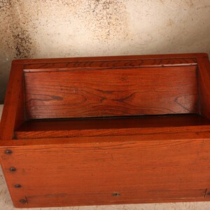 May include: A rectangular wooden box with a reddish-brown finish and visible wood grain. The box features a slanted top lid, a flat front panel with a small central keyhole, and four dark metal rivets aligned vertically along the left side edge.