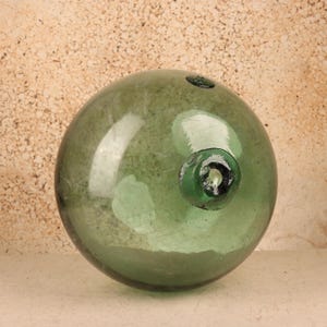 May include: A large, spherical, translucent green glass fishing float. The surface of the glass is smooth with visible internal bubbles and slight imperfections. A thick, circular glass seal is prominent on the upper right side of the sphere.