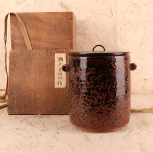 May include: A brown ceramic jar with a black lid and small handles. The jar has a speckled pattern and sits next to a wooden box with a label featuring Japanese text. A tan strap is attached to the box.